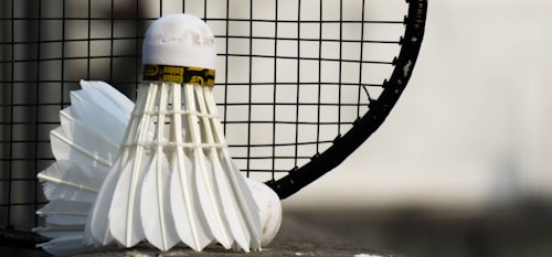 A close-up view of a shuttlecock leaning against a badminton racket with a focus on the net and feathers. The background is blurred, emphasizing the sports equipment in the foreground.