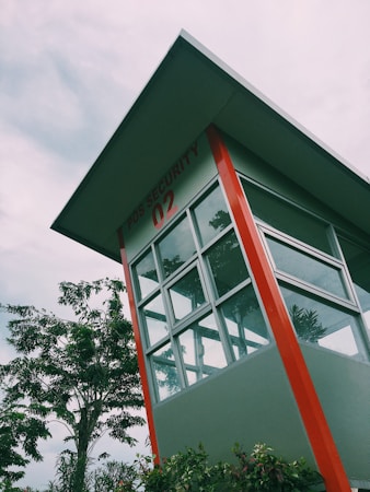 A security post with large glass windows under a slanted roof. The building is primarily grey with prominent red accents. In the background, there are lush green trees and overcast skies, suggesting a cloudy day. The post has 'POS SECURITY 02' written on it.