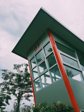A security post with large glass windows under a slanted roof. The building is primarily grey with prominent red accents. In the background, there are lush green trees and overcast skies, suggesting a cloudy day. The post has 'POS SECURITY 02' written on it.