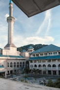 A large mosque features prominently, with an elegant minaret rising into the sky. The building has a series of arches and a blue-green roof. In the foreground, a courtyard with trees and a few people walking is visible. In the distance, a range of hills adds to the scenic backdrop, all under a clear blue sky with a few clouds.