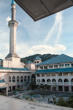 A large mosque features prominently, with an elegant minaret rising into the sky. The building has a series of arches and a blue-green roof. In the foreground, a courtyard with trees and a few people walking is visible. In the distance, a range of hills adds to the scenic backdrop, all under a clear blue sky with a few clouds.