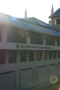 A large building with a prominent sign reading 'THE IIUM LIBRARY' and a web address. Architecture includes pointed towers which are highlighted by lens flares due to sunlight. The facade features multiple windows and patterned panels.