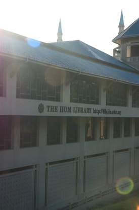 A large building with a prominent sign reading 'THE IIUM LIBRARY' and a web address. Architecture includes pointed towers which are highlighted by lens flares due to sunlight. The facade features multiple windows and patterned panels.