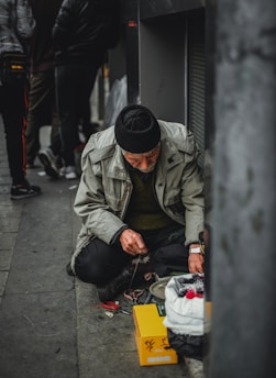 man in gray jacket and black knit cap sitting on sidewalk during daytime