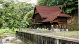 A traditional wooden house with a steep gabled roof is nestled in a lush, tropical forest. The house is elevated on stilts and features intricate wooden carvings. A small stream runs alongside the property, and the area is surrounded by dense greenery.