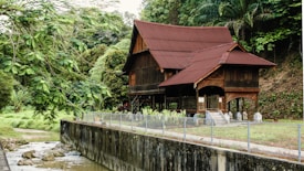 A traditional wooden house with a steep gabled roof is nestled in a lush, tropical forest. The house is elevated on stilts and features intricate wooden carvings. A small stream runs alongside the property, and the area is surrounded by dense greenery.
