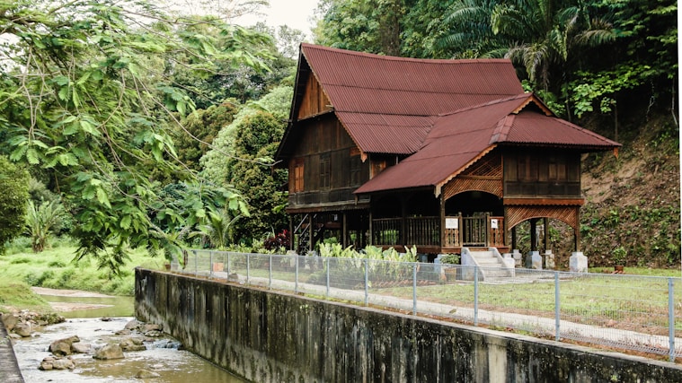 A traditional wooden house with a steep gabled roof is nestled in a lush, tropical forest. The house is elevated on stilts and features intricate wooden carvings. A small stream runs alongside the property, and the area is surrounded by dense greenery.