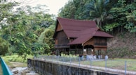 A traditional wooden house with a steep, red roof is situated amidst lush greenery. The house is elevated on stilts, featuring intricate wooden latticework and surrounded by dense foliage. A narrow stream is visible in the foreground, bordered by a concrete embankment and a metal fence.