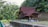 A traditional wooden house with a steep, red roof is situated amidst lush greenery. The house is elevated on stilts, featuring intricate wooden latticework and surrounded by dense foliage. A narrow stream is visible in the foreground, bordered by a concrete embankment and a metal fence.
