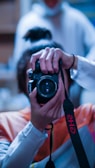A person holding a professional camera up to their face as if taking a photograph. The camera is black with a prominent lens. The person is wearing a light-colored long sleeve shirt, and their hair is tied back. The background is blurred, suggesting a shallow depth of field.