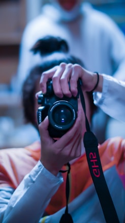 A person holding a professional camera up to their face as if taking a photograph. The camera is black with a prominent lens. The person is wearing a light-colored long sleeve shirt, and their hair is tied back. The background is blurred, suggesting a shallow depth of field.
