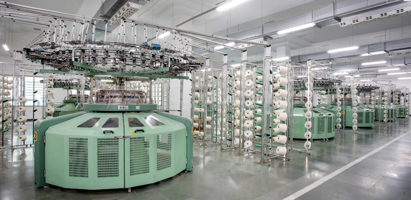 A knitted fabric machine threaded with rows of yarn on a mill floor.