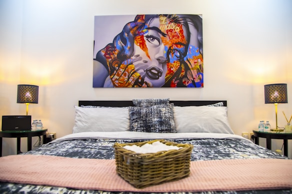 A stylish bedroom featuring a bed with patterned pillows and a blanket, flanked by two nightstands. Each nightstand has a decorative lamp with intricate patterns, and there are bottles of water placed alongside other small items. Above the bed, a large, colorful artwork of a woman's face with an abstract design adds vibrancy to the room. A woven basket, possibly holding towels, is placed at the foot of the bed.