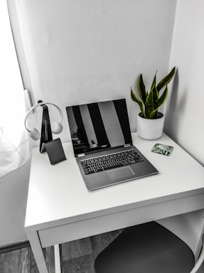 A minimalist workspace with a sleek laptop, soft green plant, and muted pink notebook on a clean white desk.