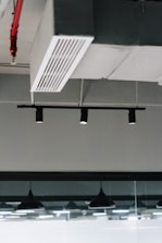 An industrial ceiling with a metal air vent and black recessed track lighting. There is a visible fire sprinkler system with red piping and modern pendant lights reflected in a glass partition.