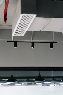 An industrial ceiling with a metal air vent and black recessed track lighting. There is a visible fire sprinkler system with red piping and modern pendant lights reflected in a glass partition.
