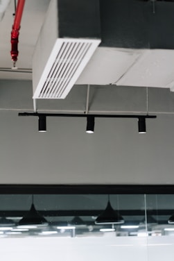 An industrial ceiling with a metal air vent and black recessed track lighting. There is a visible fire sprinkler system with red piping and modern pendant lights reflected in a glass partition.