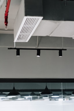 An industrial ceiling with a metal air vent and black recessed track lighting. There is a visible fire sprinkler system with red piping and modern pendant lights reflected in a glass partition.
