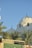 The image features two white domes with crescent moon finials, typical in Islamic architecture, surrounded by palm trees. The sky is clear blue, and there is a green and beige signage at the bottom with Arabic text.