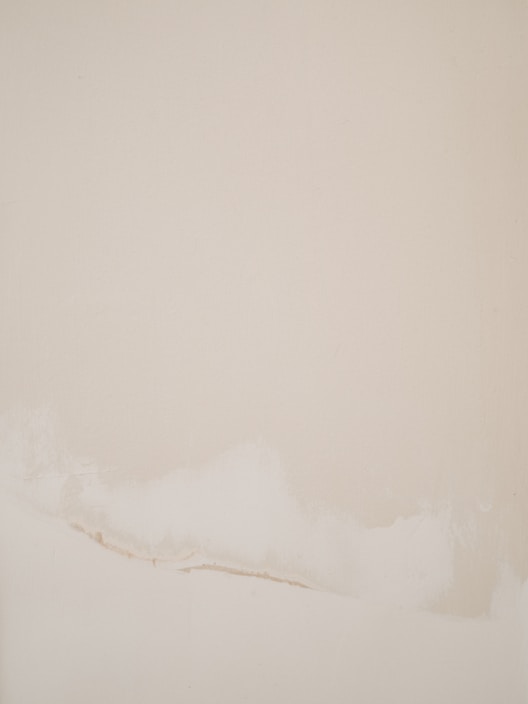A minimalist abstract painting with a soft beige background and subtle white and cream brush strokes along the lower portion.