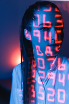 A person with long hair is standing in a dimly lit environment. Bright red digital numbers are projected across their face, creating a futuristic and technological effect. The background features blue tones with a warm orange light source adding contrast.