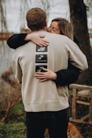A couple embraces warmly outdoors, with the woman hugging a man who has ultrasound photos attached to the back of his sweatshirt. They're near a body of water, surrounded by natural elements like grass and trees.
