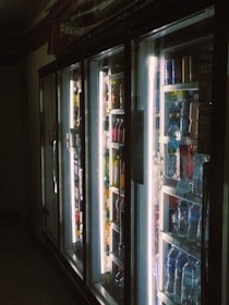 A dimly lit aisle in a store featuring a row of refrigerator doors. Behind the glass, various bottled beverages are visible, including water and soda. The interior lights of the refrigerators create a stark contrast against the darker surroundings, highlighting the colorful array of bottles.