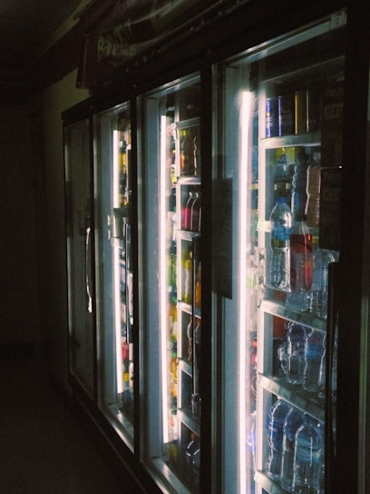 A dimly lit aisle in a store featuring a row of refrigerator doors. Behind the glass, various bottled beverages are visible, including water and soda. The interior lights of the refrigerators create a stark contrast against the darker surroundings, highlighting the colorful array of bottles.