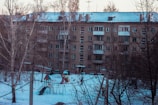 A residential apartment building with multiple balconies covered in snow. The foreground shows a snow-covered playground with swings and a slide surrounded by leafless trees.