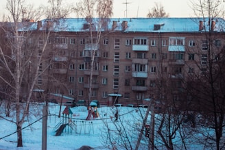 A residential apartment building with multiple balconies covered in snow. The foreground shows a snow-covered playground with swings and a slide surrounded by leafless trees.
