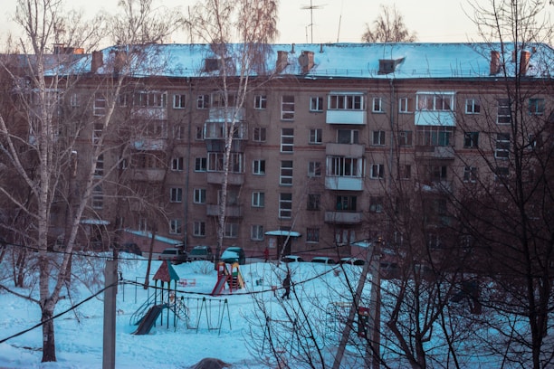 A residential apartment building with multiple balconies covered in snow. The foreground shows a snow-covered playground with swings and a slide surrounded by leafless trees.