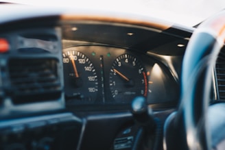 A car dashboard featuring speedometer and tachometer gauges, with the speedometer showing speeds up to 180 and the tachometer indicating revolutions per minute. The foreground includes parts of the steering wheel, and the dashboard appears to be in a vehicle interior setting.