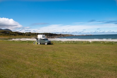 A serene coastal landscape featuring a grassy field leading to a sandy beach with gentle waves. A camper vehicle with a rooftop tent is parked on the grass, indicating a peaceful camping spot. The background reveals rocky outcrops and a mix of blue sky with scattered clouds.