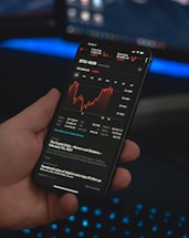 A person holds a smartphone displaying a financial application with cryptocurrency statistics and graphs, specifically showing BTC-EUR rates. The screen is predominantly dark with red and white text and graphs. The background features a blurred computer setup with a keyboard emitting a blue glow.