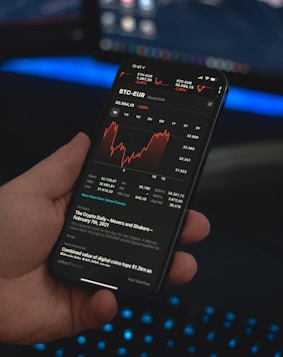 A person holds a smartphone displaying a financial application with cryptocurrency statistics and graphs, specifically showing BTC-EUR rates. The screen is predominantly dark with red and white text and graphs. The background features a blurred computer setup with a keyboard emitting a blue glow.