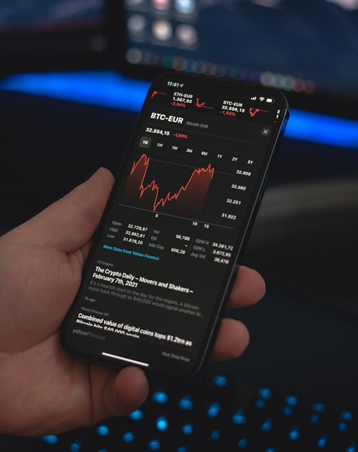 A person holds a smartphone displaying a financial application with cryptocurrency statistics and graphs, specifically showing BTC-EUR rates. The screen is predominantly dark with red and white text and graphs. The background features a blurred computer setup with a keyboard emitting a blue glow.