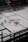 A group of ice hockey players in red uniforms practice on an indoor rink, surrounded by empty stands. The ice features a large central logo and lines for gameplay. Coaches or staff appear to be on the sidelines, while a few players in white jerseys skate across the rink.