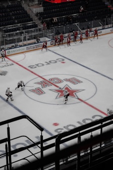 A group of ice hockey players in red uniforms practice on an indoor rink, surrounded by empty stands. The ice features a large central logo and lines for gameplay. Coaches or staff appear to be on the sidelines, while a few players in white jerseys skate across the rink.