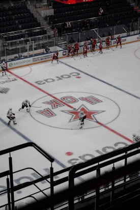 A group of ice hockey players in red uniforms practice on an indoor rink, surrounded by empty stands. The ice features a large central logo and lines for gameplay. Coaches or staff appear to be on the sidelines, while a few players in white jerseys skate across the rink.