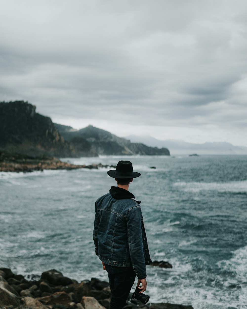 man in blue denim jacket standing on rock near body of water during daytime