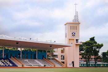A sports field with a large seating area that includes blue and white bleachers. A prominent watchtower with a cross on top stands next to the seating area, displaying a crest. The area is surrounded by trees and has a cloudy sky overhead.