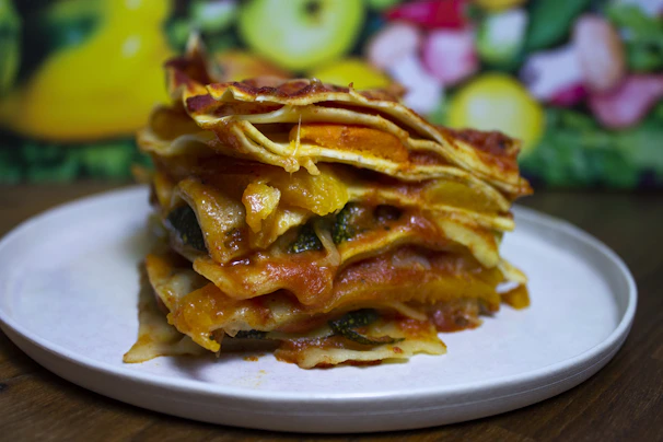A close-up of a colorful vegetable lasagna stack on a white plate, featuring layers of pasta, red sauce, yellow bell peppers, and green zucchini with a blurred background of vibrant fruits and vegetables.