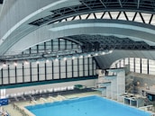 An indoor aquatic center features a large swimming pool with multiple diving platforms. The curved, modern architectural ceiling and large window panels provide ample lighting and a spacious atmosphere. Seating areas and equipment are visible around the pool area.