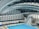 An indoor aquatic center features a large swimming pool with multiple diving platforms. The curved, modern architectural ceiling and large window panels provide ample lighting and a spacious atmosphere. Seating areas and equipment are visible around the pool area.