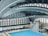 An indoor aquatic center features a large swimming pool with multiple diving platforms. The curved, modern architectural ceiling and large window panels provide ample lighting and a spacious atmosphere. Seating areas and equipment are visible around the pool area.