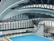 An indoor aquatic center features a large swimming pool with multiple diving platforms. The curved, modern architectural ceiling and large window panels provide ample lighting and a spacious atmosphere. Seating areas and equipment are visible around the pool area.