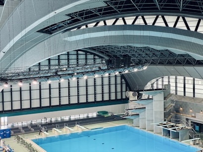 An indoor aquatic center features a large swimming pool with multiple diving platforms. The curved, modern architectural ceiling and large window panels provide ample lighting and a spacious atmosphere. Seating areas and equipment are visible around the pool area.