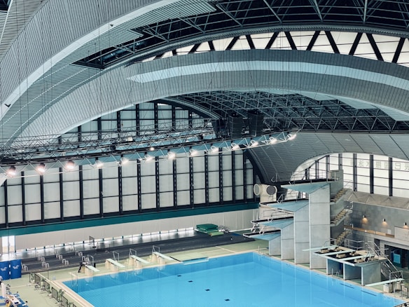 An indoor aquatic center features a large swimming pool with multiple diving platforms. The curved, modern architectural ceiling and large window panels provide ample lighting and a spacious atmosphere. Seating areas and equipment are visible around the pool area.