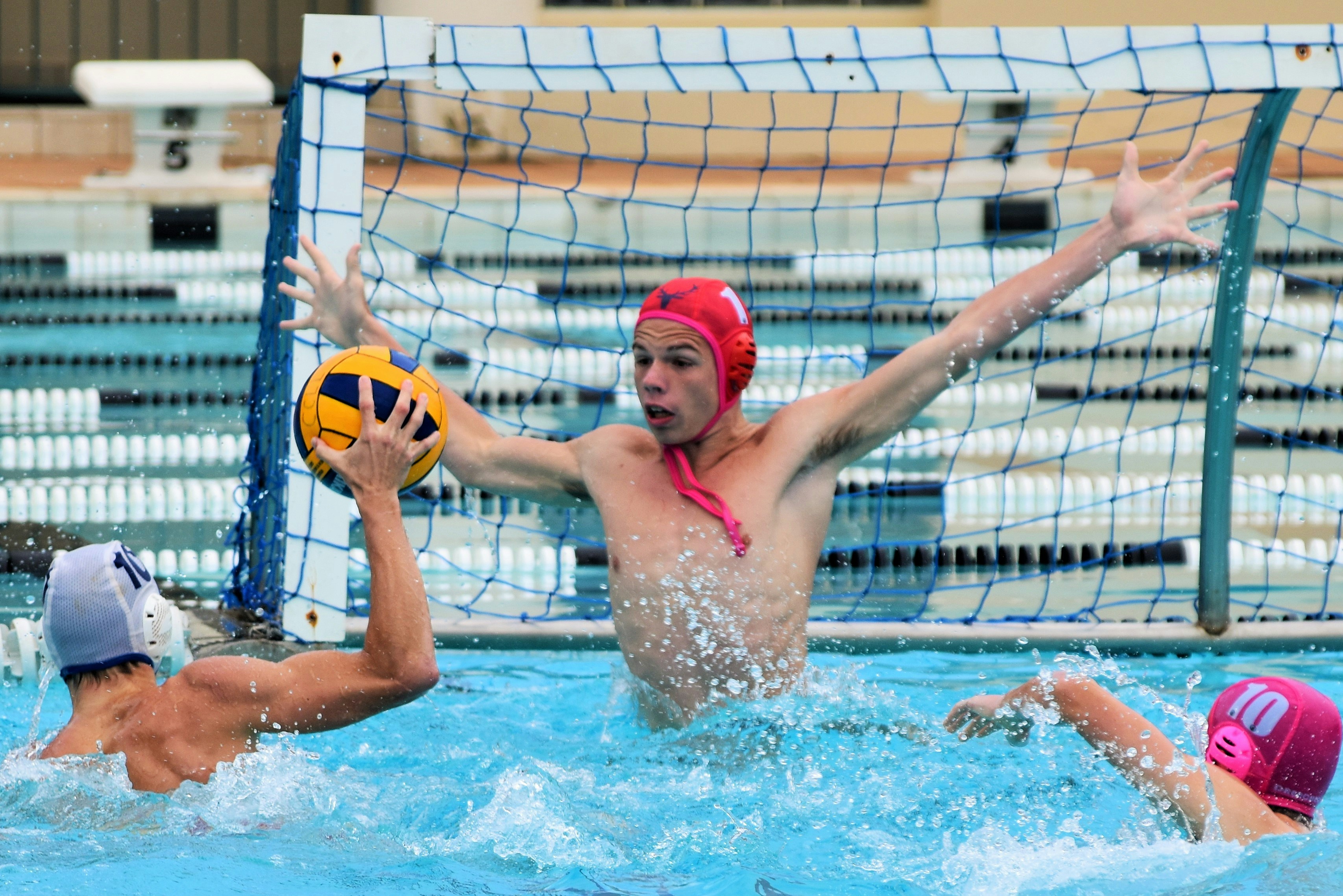 During a water polo match the goal keeper (red cap) lifts himself out of the water and spreads his arms as wide as possible to prevent the other team scoring.