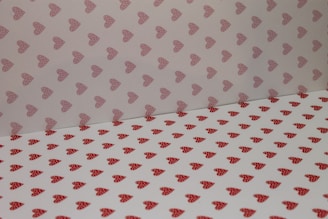 A pattern of heart shapes covers a surface and wall. The hearts are red with polka dots on a white background. The upper section has a lighter shade with similar heart patterns.